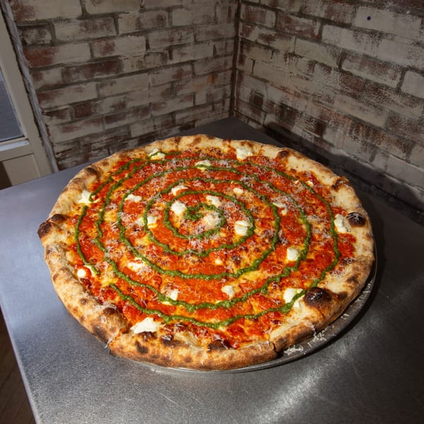 Jonny's Pizza with red sauce, white cheese, green pesto swirl, and grated cheese on a metal table.