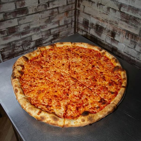 A whole Jonny's Pizza cheese pizza, cut into slices, on a metal table against a brick wall.